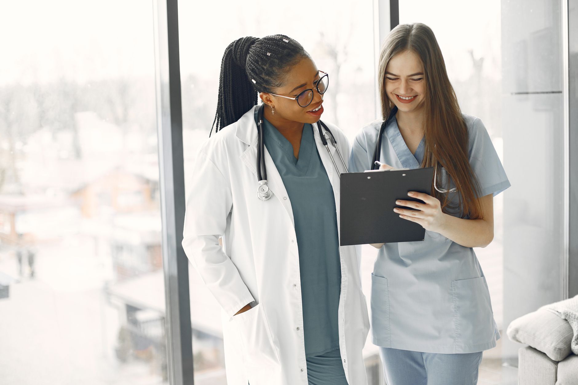 Two healthcare workers, one in a white coat and one in blue scrubs, reviewing a document on a clipboard together beside a bright window